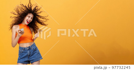 Portrait of joiyful carefree and happy charming european woman with curly hairstyle laughing out loud holding smartphone wearing wireless earbuds listening awesome music via app over orange wall 97732245
