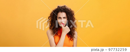 Waist-up shot of serious determined and focused displeased woman with curly hairstyle frowning biting thumb looking dangerous and angry standing daring and rebellious over orange background Waist-up shot of serious determined and focused displeased woman with curly hairstyle frowning biting thumb looking dangerous and angry standing daring and rebellious over orange background 97732259
