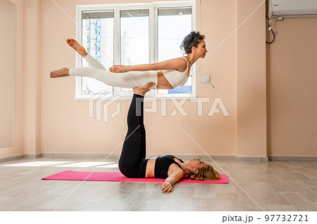 Steam yoga of two women doing yoga indoors. One woman holding the other with her arms and legs 97732721