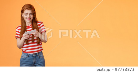 Excited redhead caucasian girl look intense, frowning, trying pass hard game level, playing new arcade, tap screen, hold smartphone horizontal, look telephone display cautious, orange background 97733873