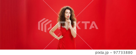 Shh let us keep it between us. Attractive and elegant adult successful woman in stylish evening dress shushing with index finger over mouth hiding secret or making surprise over red background 97733880