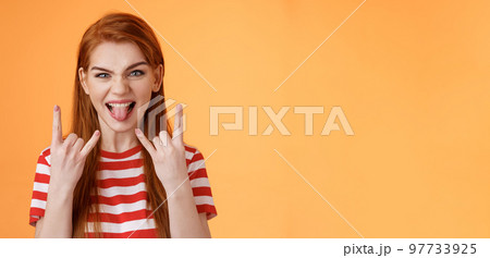 Lets rock-n-roll. Daring cheerful good-looking caucasian redhead woman, having fun, show heavy metal gestures, stick tongue and smile amused, enjoy music festival, stand orange background Lets rock-n-roll. Daring cheerful good-looking caucasian redhead woman, having fun, show heavy metal gestures, stick tongue and smile amused, enjoy music festival, stand orange background 97733925