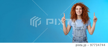 Motivated happy cheerful redhead silly curly woman pointing up inrdex finger smiling charmed impressed excited showing awesome promo discuss interesting advertisement blue background Motivated happy cheerful redhead silly curly woman pointing up inrdex finger smiling charmed impressed excited showing awesome promo discuss interesting advertisement blue background 97734744