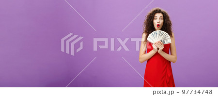 Studio shot of woman being lucky in casino winning lots of cash holding money in hands saying wow with folded lips staring impressed and excited over purple wall in evening red dress Studio shot of woman being lucky in casino winning lots of cash holding money in hands saying wow with folded lips staring impressed and excited over purple wall in evening red dress 97734748