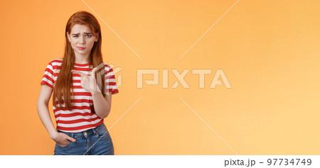 Upset uneasy cute ginger gitl long natural red hair, frowning smirking hesitant, complain, looking suspicious and doubtful point left, not sure if product really good, feel unsure, orange background 97734749
