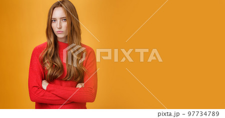 Suspicious intense and defensive ginger girl standing in passive-aggressive pose pouting and frowning looking with disbelief and disdain at camera, offended against orange background 97734789