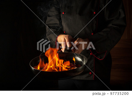 Professional chef cooking food in pan with fire flame on black background. Restaurant and hotel service concept. European cuisine Professional chef cooking food in pan with fire flame on black background. Restaurant and hotel service concept. European cuisine 97735200