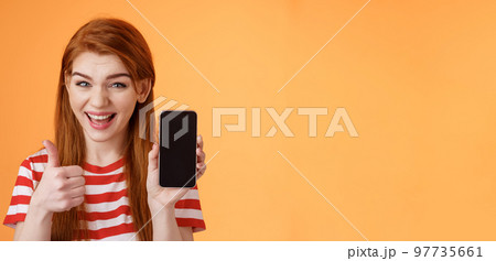 Cheerful assertive redhead woman recommend smartphone app, show phone display, thumb-up like approval sign, smiling broadly, suggest good online purchase, mobile advertisement Cheerful assertive redhead woman recommend smartphone app, show phone display, thumb-up like approval sign, smiling broadly, suggest good online purchase, mobile advertisement 97735661