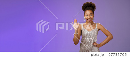Friendly elegant african-american young 20s woman wearing elegant silver glittering dress hold hand waist waving hello greeting gesture say hi informally meet friend talking blue background Friendly elegant african-american young 20s woman wearing elegant silver glittering dress hold hand waist waving hello greeting gesture say hi informally meet friend talking blue background 97735706