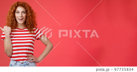 Sassy good-looking emotive happy smiling redhead european female curly hairstyle pointing thumb left grin assertive cheeky hold hand waist directing promo advertising offer red background Sassy good-looking emotive happy smiling redhead european female curly hairstyle pointing thumb left grin assertive cheeky hold hand waist directing promo advertising offer red background 97735834