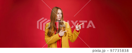 Studio shot of intense suspicious and displeased redhead female in yellow coat pointing and looking serious left squnting and frowning making focused look at something disturbing and doubtful 97735838