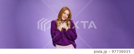 How cute thanks. Tender charming redhead girl in purple sweater holding hands on heart tilting head flirty and happy as smiling at camera thankful and touched with romantic gift over violet wall 97736035