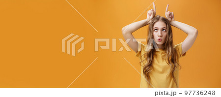 GIrl going crazy from boredom making freaky funny faces fooling around squinting holding index fingers on head like horns sticking out tongue and folding lips aping over orange background 97736248