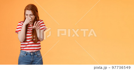 Gosh it stinks. Reluctant disgusted redhead woman raise hand block dislike gesture shut nose, cringe from aversion dislike, puke from reek bad smell, stand orange background disappointed 97736549