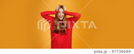 Excited and overwhelmed charming redhead woman in knitted warm red dress yelling from thrill and happiness holding hands on hair smiling broadly impressed with unexpected luck over orange wall Excited and overwhelmed charming redhead woman in knitted warm red dress yelling from thrill and happiness holding hands on hair smiling broadly impressed with unexpected luck over orange wall 97736689