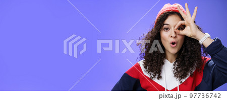Amused girl having fun showing okay or zero gesture over eye folding lips from amazement as looking and surprising curious thing, standing impressed and delighted over blue wall in winter hat 97736742