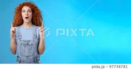 Amused astonished impressionable cute redhead curly girl open mouth drop jaw gasping excited stare camera thrilled pointing index fingers up thrilled awesome store offer sale blue background 97736781