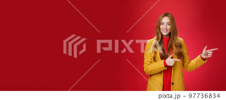 Carefree cute and tender young redhead female student in stylish yellow coat over dress pointing right and smiling with delighted and impressed look showing great place over red background 97736834