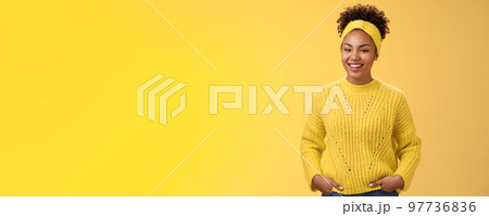 Friendly outgoing relaxed young african-american beautiful girl headband forehead in sweater hold hands pockets smiling broadly have fun talking interesting communication, standing yellow wall 97736836