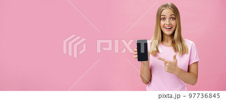 Cheerful attractive and pleasant woman promoting cool app or smartphone holding cellphone and pointing at device screen smiling amused and impressed standing over pink background Cheerful attractive and pleasant woman promoting cool app or smartphone holding cellphone and pointing at device screen smiling amused and impressed standing over pink background 97736845