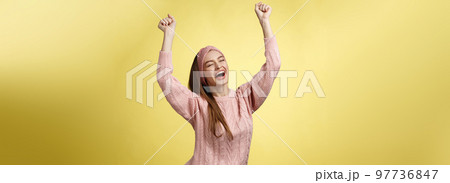 Positive celebating happy european young woman wearing casual sweater yelling happily shouting yes, triumphing racing hands up in success jumping ecstatic rejoicing cheerfully over success 97736847