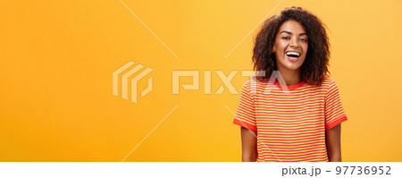 Waist-up shot of outgoing happy charming dark-skinned female with curly hairstyle laughing joyfully posing in striped trendy t-shirt over orange background enjoying nice casual conversation Waist-up shot of outgoing happy charming dark-skinned female with curly hairstyle laughing joyfully posing in striped trendy t-shirt over orange background enjoying nice casual conversation 97736952