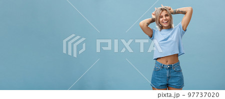 Studio shot of cute attractive and joyful stylish blond woman with tattoos on arms winking and smiling broadly holding index fingers on head making horns standing in summer outfit over blue wall Studio shot of cute attractive and joyful stylish blond woman with tattoos on arms winking and smiling broadly holding index fingers on head making horns standing in summer outfit over blue wall 97737020