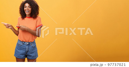 Ask him. Portrait of friendly and joyful good-looking stylish female shop assistant with curly hair and dark skin pointing left with both hands, smiling assured and entertained over orange background Ask him. Portrait of friendly and joyful good-looking stylish female shop assistant with curly hair and dark skin pointing left with both hands, smiling assured and entertained over orange background 97737021