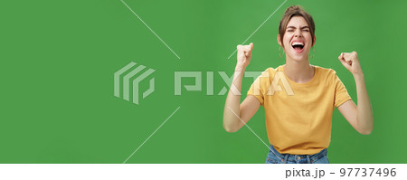 Waist-up shot of charismatic energized and excited female in yellow t-shirt closing eyes yelling from joy and happiness raising hands in cheer celebrating successful news over green background 97737496