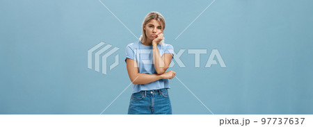 Studio shot of displeased moody girlfriend feeling disappointed boyfriend spoilt date leaning head on palm sulking from dissatisfaction and squinting being offended posing over blue background 97737637