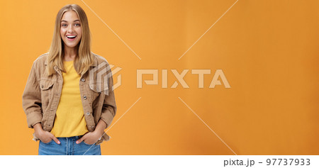 Charismatic tanned woman in corduroy jacket and yellow t-shirt ready for chilly autumn walk with friends smiling joyfully gazing entertained at camera holding hand in pockets casually over orange wall 97737933