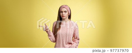 Pissed puzzled young attractive arrogant moody girlfriend in sweater, headband looking irritated, intense frowning pointing finger at copy space, directing person sits on her place, annoyed 97737970