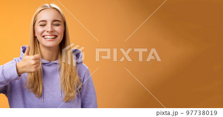 Amused joyful optimistic young excited blond european female model in hoodie smiling broadly close eyes having fun show approval gesture thumbs-up like awesome trip vacation idea, orange background 97738019