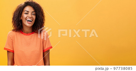 Waist-up shot of outgoing happy charming dark-skinned female with curly hairstyle laughing joyfully posing in striped trendy t-shirt over orange background enjoying nice casual conversation 97738085