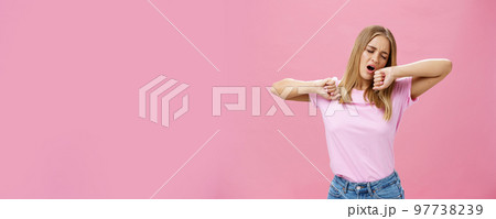 Too tired to work today. Lazy and exhausted attractive young female student doing homework all night yawning with closed eyes while stretching hands from after tiresome project over pink wall 97738239