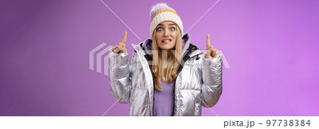Scared unsure worried awkward young cute blond girl biting lip stooping pointing up widen eyes nervously scared someone find out secret standing embarrassed and intense purple background 97738384