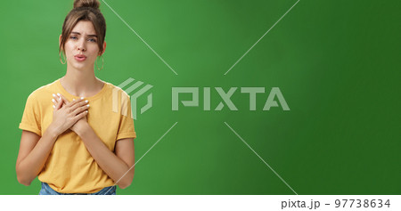 Touched and pleased attractive young woman in yellow t-shirt holding palms pressed to chest in thankful and grateful gesture folding lips melting heart seeing cute thing over green background 97738634