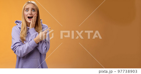 Amazed excited overwhelmed young blond girl in hoodie press palm cheek screaming astonished pointing right seeing famous super star cannot hold joyful positive emotions, orange background 97738903