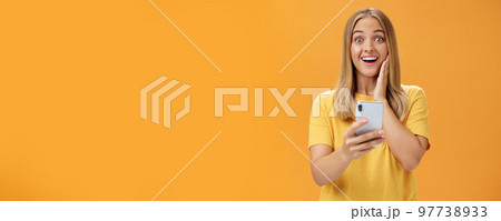 Portrait of surprised and impressed woman reacting to awesome app in smartphone touching cheek from amazement and joy smiling broadly at camera holding cellphone in hand over orange wall Portrait of surprised and impressed woman reacting to awesome app in smartphone touching cheek from amazement and joy smiling broadly at camera holding cellphone in hand over orange wall 97738933