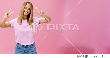 Cute chubby carefree young woman with short fair hair tilting head showing peace gestures with happy friendly smile having fun spending time amused and joyful against pink background 97739159