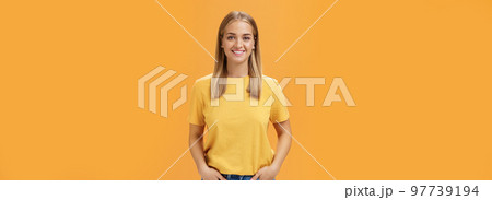 Cute chubby female with tanned skin and fair hair posing optimistic and joyful against orange background holding hands in pockets smiling broadly at camera charismatic and friendly-looking 97739194