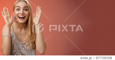 Impressed charming entertained blond european 25s woman in stylish silver glamour dress having fun playing peekaboo smiling joyfully hold palms near face laugh amused, standing red background 97739206