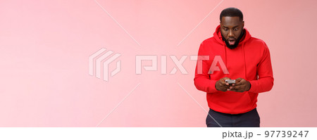 Amused focused impressed african-american bearded guy in red hoodie drop jaw widen eyes look amazed smartphone display winning hard game level answering exciting message, standing pink background Amused focused impressed african-american bearded guy in red hoodie drop jaw widen eyes look amazed smartphone display winning hard game level answering exciting message, standing pink background 97739247