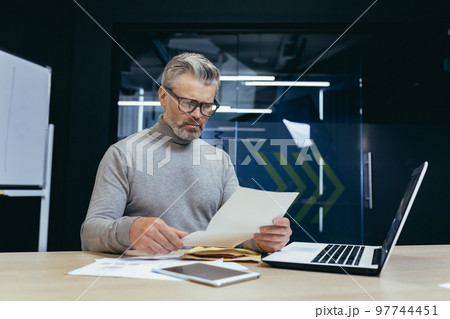 Mature gray-haired businessman inside modern office received letter message, senior man upset opens postal envelope, boss reads bad news upset and sad working at desk with laptop. Mature gray-haired businessman inside modern office received letter message, senior man upset opens postal envelope, boss reads bad news upset and sad working at desk with laptop. 97744451