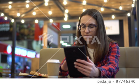 A young woman eats pizza in a cafe and uses a tablet. The girl is reading a book on a tablet. The cafe is cozy decorated with light bulbs. 97746340