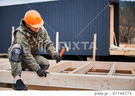 Man worker building wooden frame house. Carpenter hammering nail into wooden board, using hammer. Carpentry concept. Man worker building wooden frame house. Carpenter hammering nail into wooden board, using hammer. Carpentry concept. 97746773