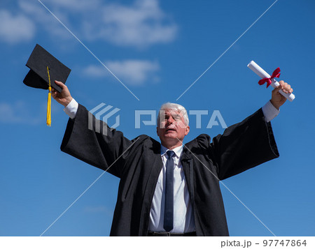 Elderly male graduate rejoices in receiving a diploma against a blue sky. Elderly male graduate rejoices in receiving a diploma against a blue sky. 97747864