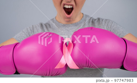 Caucasian woman in pink boxing gloves with a pink ribbon on her chest on a gray background. Fight against breast cancer. Caucasian woman in pink boxing gloves with a pink ribbon on her chest on a gray background. Fight against breast cancer. 97747983