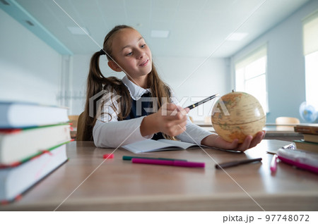 Caucasian schoolgirl sits at her desk at school and studies the globe.  97748072