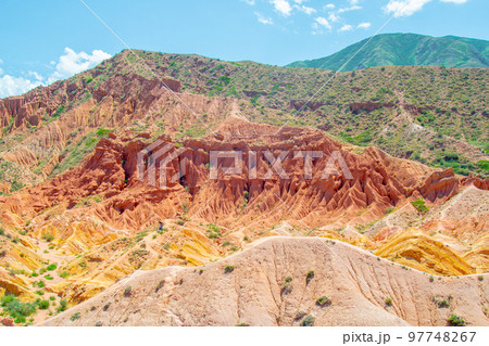 Sharp Mountain is a rock made of red orange stone and clay against a bright blue sky without clouds and desert vegetation in Fairy Tale Canyon Kyrgyzstan, Skazka Canyon. 97748267